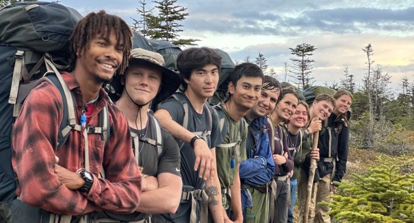 a group of gap year students wearing backpacks stand in a line and smile at the camera in a green area with trees